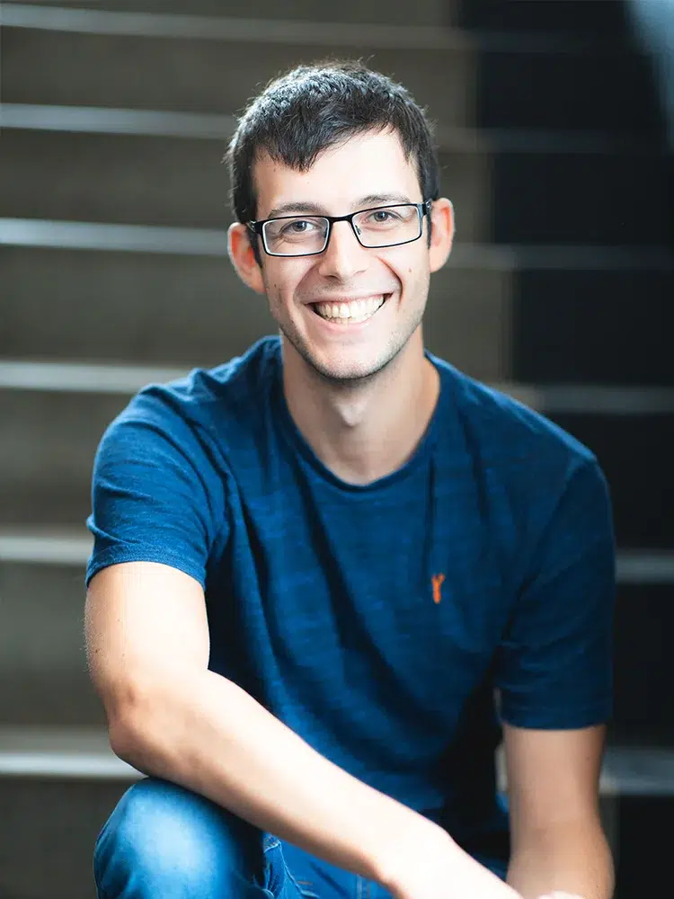 A young man, wearing glasses and a blue shirt, sits on stairs smiling.