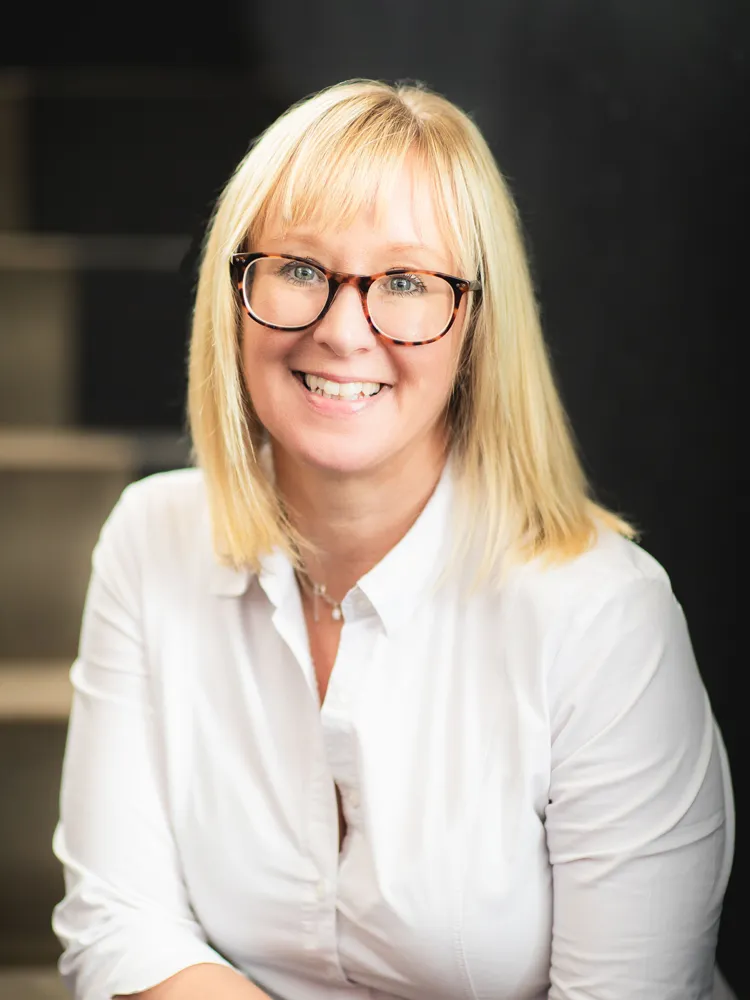 A blonde woman, wearing glasses and a white shirt, smiles at the camera in front of a dark background.