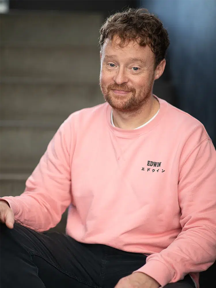 A man sits on stairs, wearing a pink Edwin sweatshirt and dark trousers, smiling at the camera.  EDWIN エドウィン is embroidered on the sweatshirt.